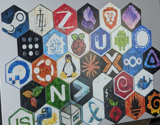 A honeycomb arrangement of hexagonal stickers on a macbook, each featuring a mosaic-style design of technology and open source logos. Visible designs include Steam, Bash, Ubuntu, Home Assistant, Raspberry Pi, Android, Tux the Linux penguin, Nextcloud, Debian, and many others. Each sticker has a textured, painterly appearance with triangular mosaic cells creating the images.